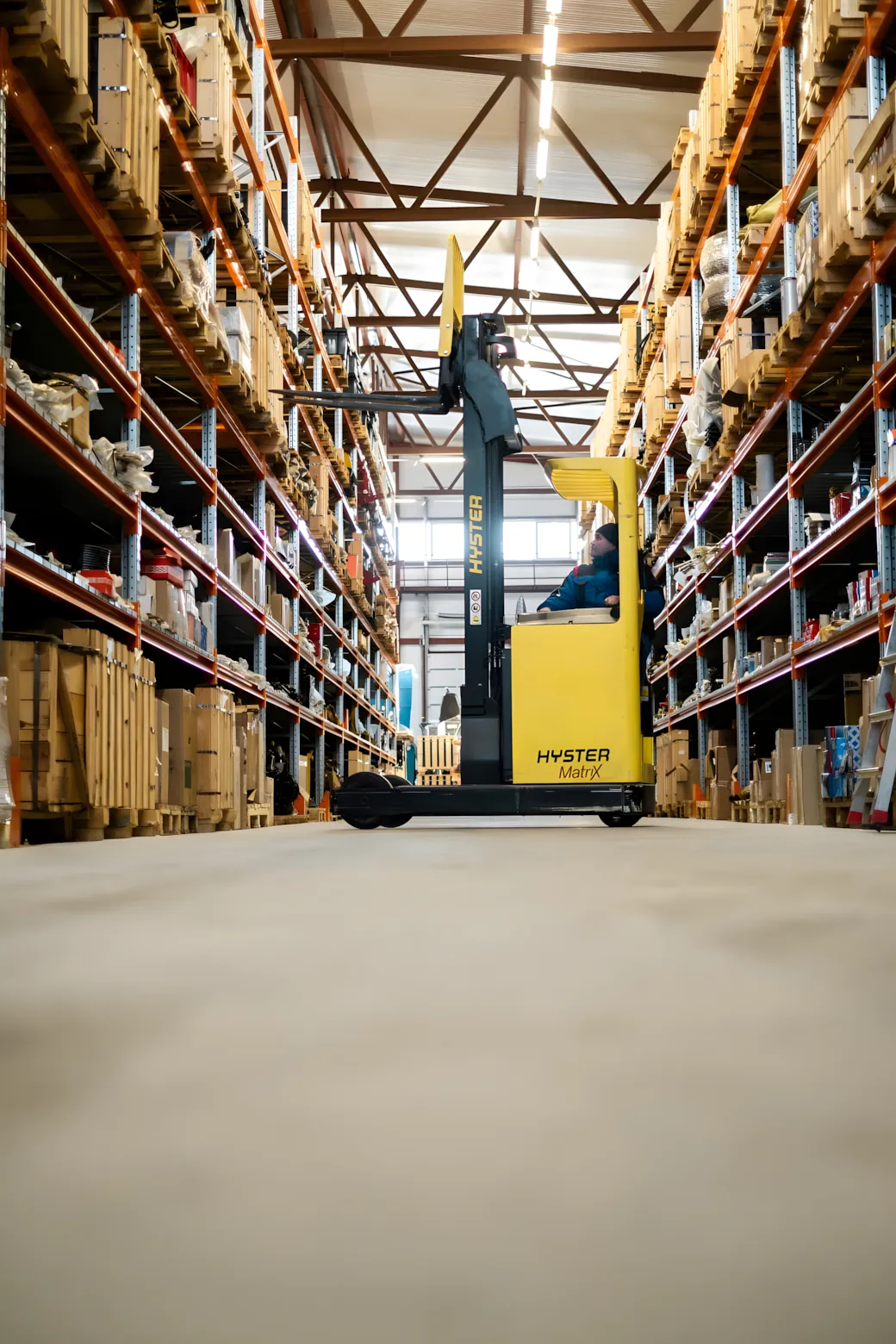 A reach truck with its mast raised in a warehouse aisle, between tall racking stacked with goods.